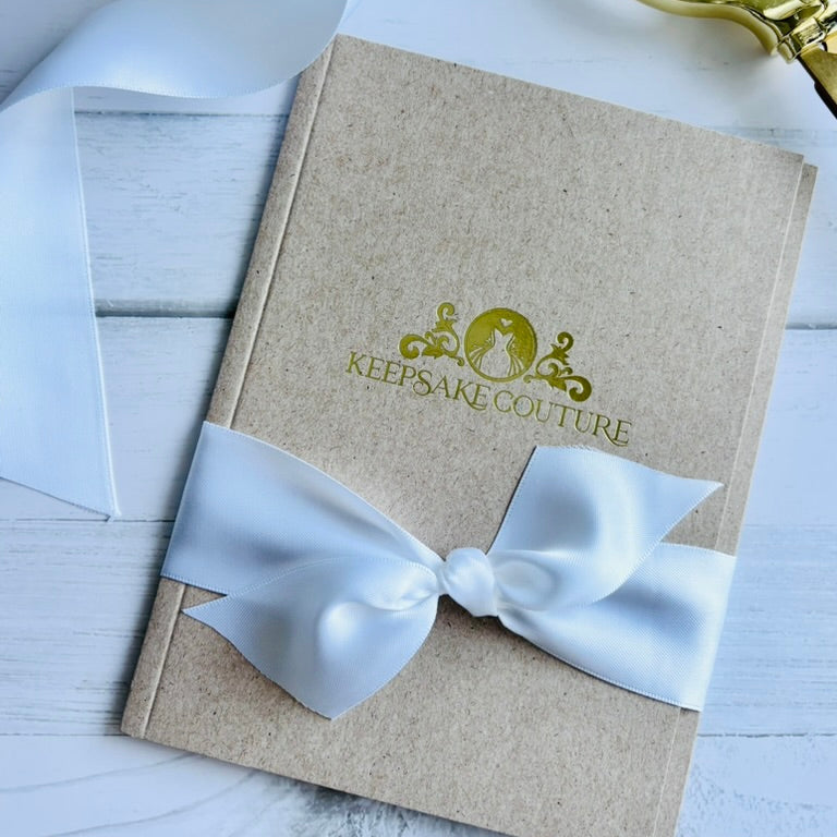 Beige notebook with a white ribbon and 'Keepsake Couture' branding on a light wooden surface.