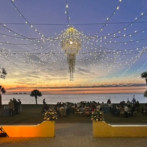 Decorative setup with string lights and a chandelier over a seated area at sunset.