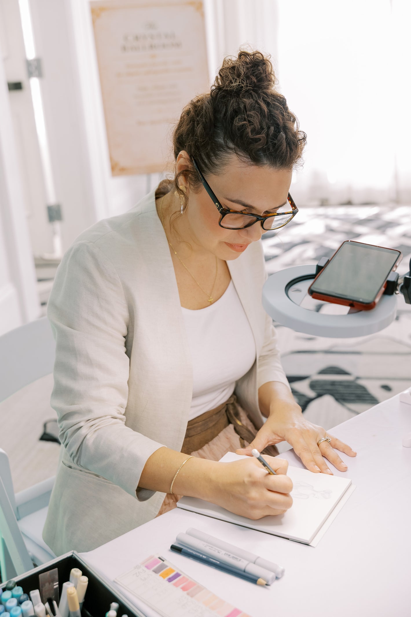 Woman sitting at a desk sketching on paper, wearing glasses.